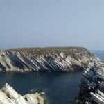 Vista de uma ilha rochosa no mar, com camadas de rocha inclinadas bem visíveis. Em primeiro plano, afloramentos rochosos enquadram uma pequena enseada.