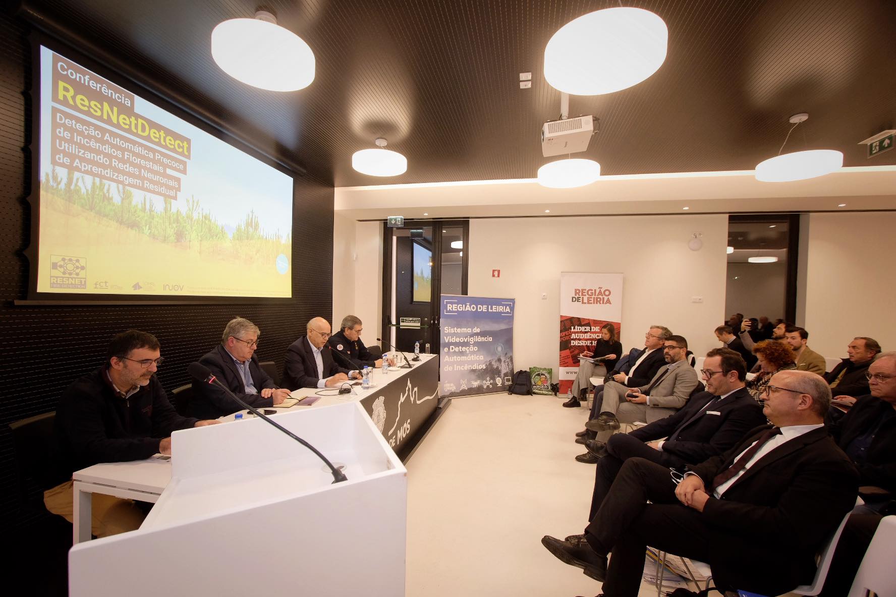 Conferência ResNetDetect sobre deteção automática precoce de incêndios florestais com recurso a redes neuronais. Vários intervenientes estão sentados numa mesa perante uma audiência, numa sala com materiais da Região de Leiria.