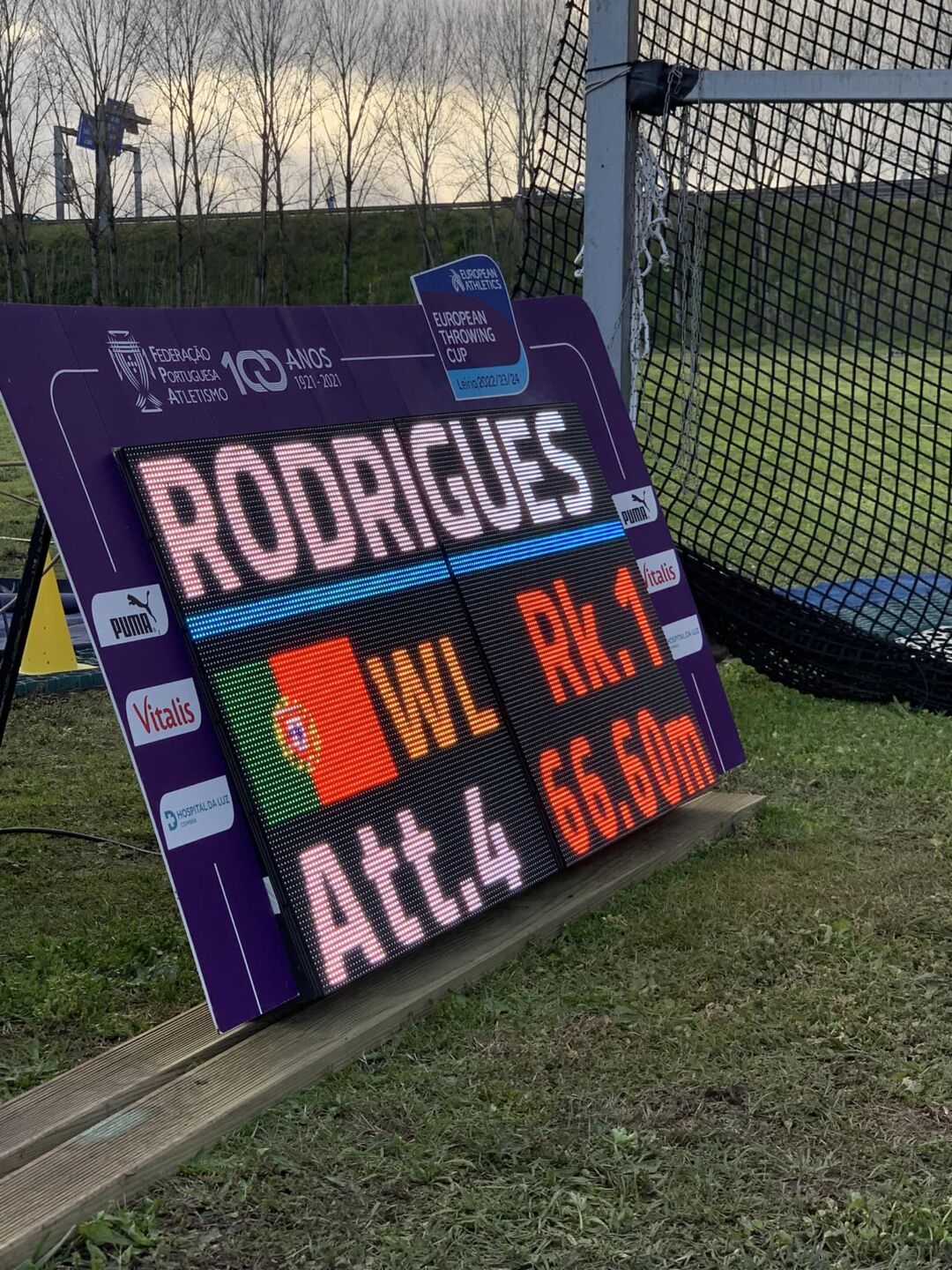 Um painel eletrónico num recinto de atletismo apresenta a indicação «RODRIGUES», a bandeira de Portugal e informações de uma prova de lançamento. Ao fundo, vê-se a rede de proteção da zona de lançamentos.