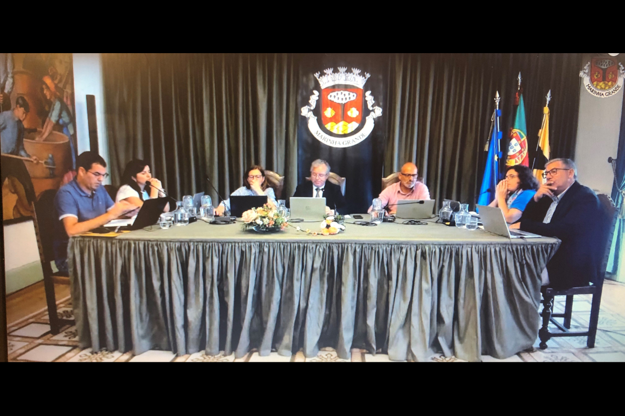 Seis pessoas estão sentadas à mesa numa reunião, com computadores portáteis e microfones. Ao fundo veem-se o brasão de Marinha Grande e bandeiras.