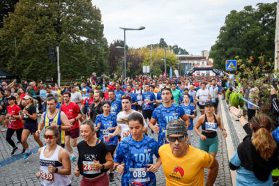 Uma multidão de participantes corre numa prova de estrada, com dorsais e camisolas de corrida, numa avenida arborizada. Há espectadores junto ao percurso.