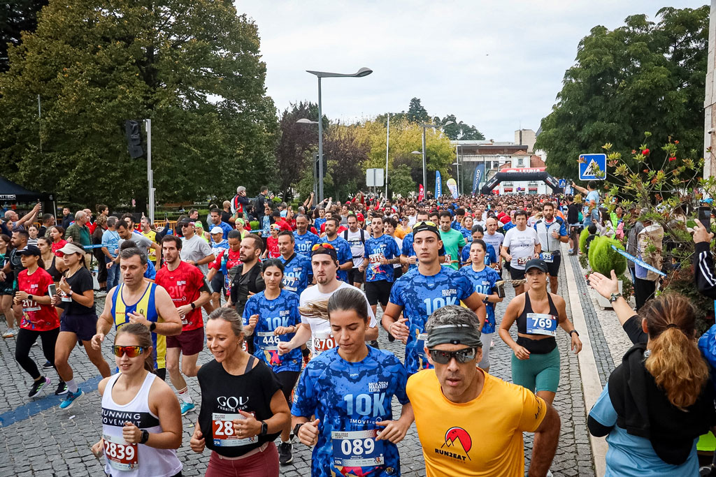Uma multidão de participantes corre numa prova de estrada, com dorsais e camisolas de corrida, numa avenida arborizada. Há espectadores junto ao percurso.