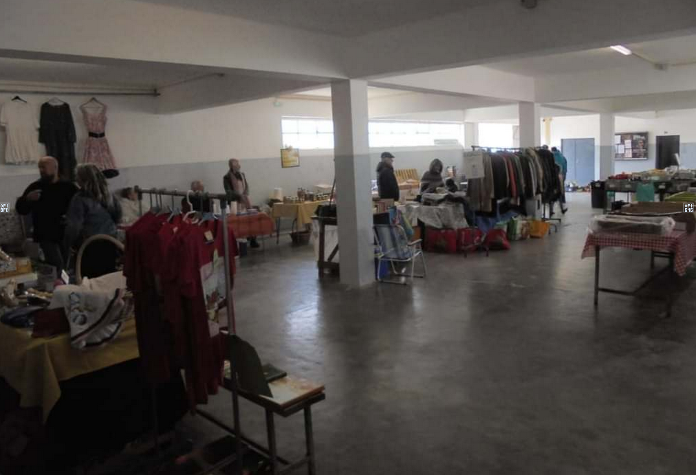 Interior de um espaço com bancas e cabides de roupa, onde várias pessoas circulam entre os artigos expostos. A disposição sugere uma feira de venda de artigos usados.
