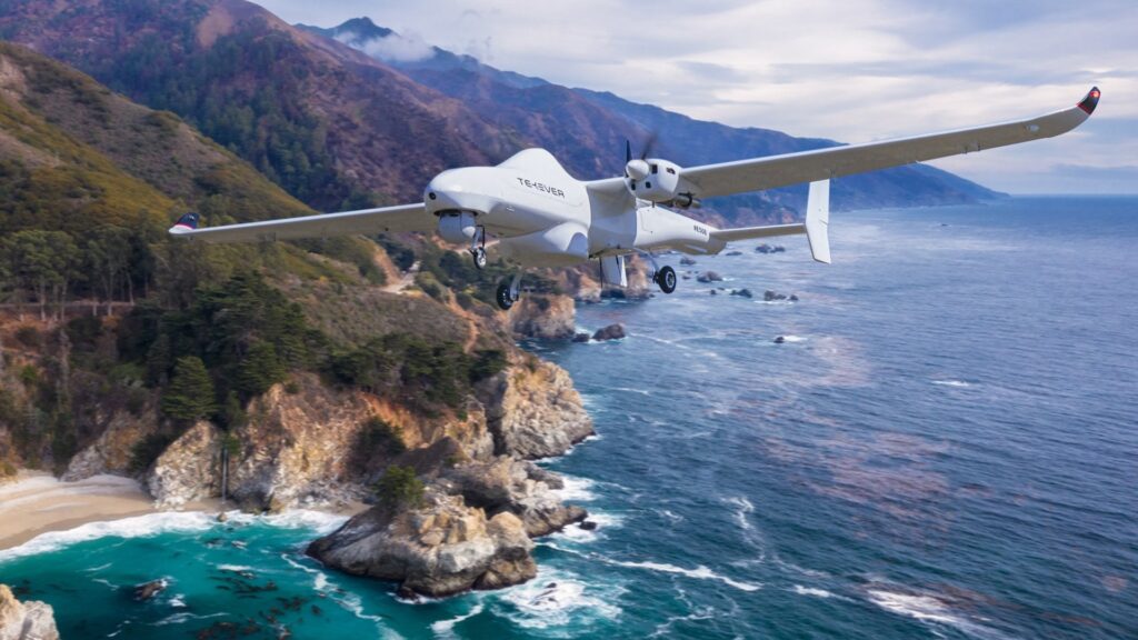 Um drone branco com a inscrição «TEKEVER» voa sobre uma costa rochosa, com o mar e montanhas ao fundo. Não há pessoas visíveis.