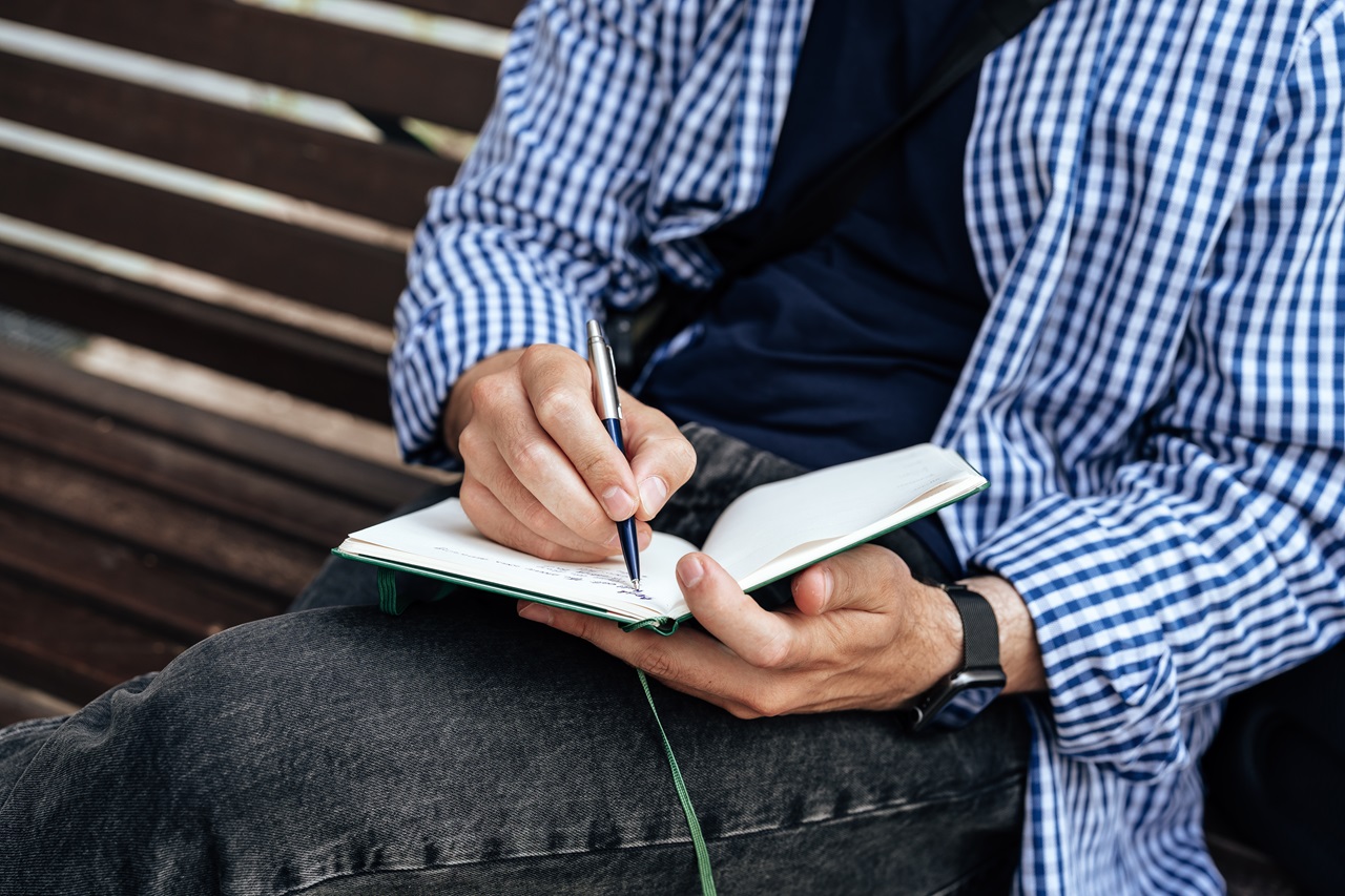 Um homem escreve num caderno enquanto está sentado no banco de um parque