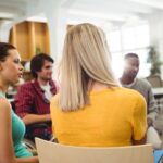 Um grupo de pessoas está sentado em círculo numa sala luminosa, aparentemente em conversa. Uma pessoa segura uma prancheta.