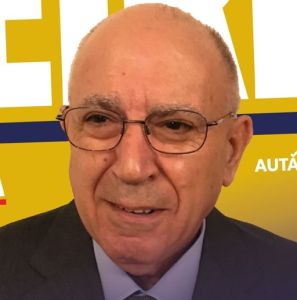 Retrato de um homem de óculos e fato escuro, diante de um fundo amarelo com letras parcialmente visíveis. A imagem não contém texto suficiente para identificar a pessoa.