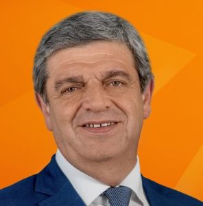 Retrato de um homem de cabelo grisalho, com fato azul e gravata, sobre um fundo laranja. Não há texto visível que permita identificá-lo.