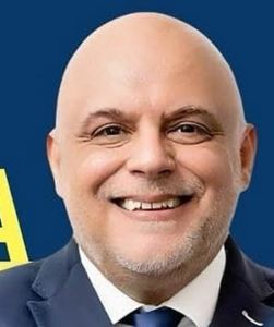Retrato de um homem careca e sorridente, vestido com fato escuro, camisola branca e gravata. O fundo é azul, com parte de uma letra amarela visível.