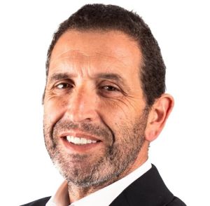 Retrato de um homem sorridente, com cabelo curto e barba, vestido com um casaco escuro e camisola branca. O fundo é branco.