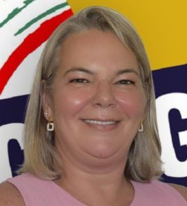 Retrato de uma mulher sorridente, de cabelo louro, com brincos e roupa cor-de-rosa. Ao fundo, vê-se um painel colorido com elementos gráficos e letras parcialmente visíveis.