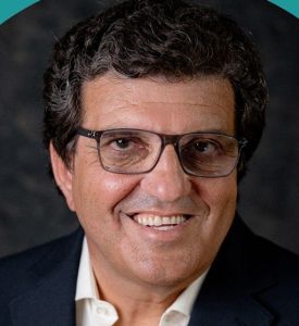 Retrato de um homem sorridente, de óculos, com camisola branca e casaco escuro. O fundo é escuro.