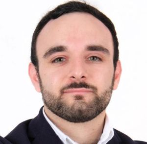 Retrato de um homem com cabelo curto escuro e barba, a olhar para a câmara. Veste um casaco escuro sobre uma camisola branca, diante de um fundo branco.