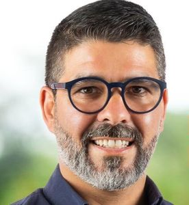 Retrato aproximado de um homem a sorrir, com óculos de armação escura e barba grisalha. O fundo está desfocado.