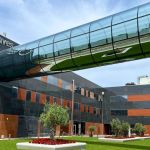 Vista exterior de um edifício com a inscrição «Lusíaves grupo». Uma passagem envidraçada elevada atravessa o espaço sobre uma zona ajardinada.