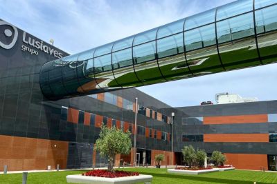 Vista exterior de um edifício com a inscrição «Lusíaves grupo». Uma passagem envidraçada elevada atravessa o espaço sobre uma zona ajardinada.