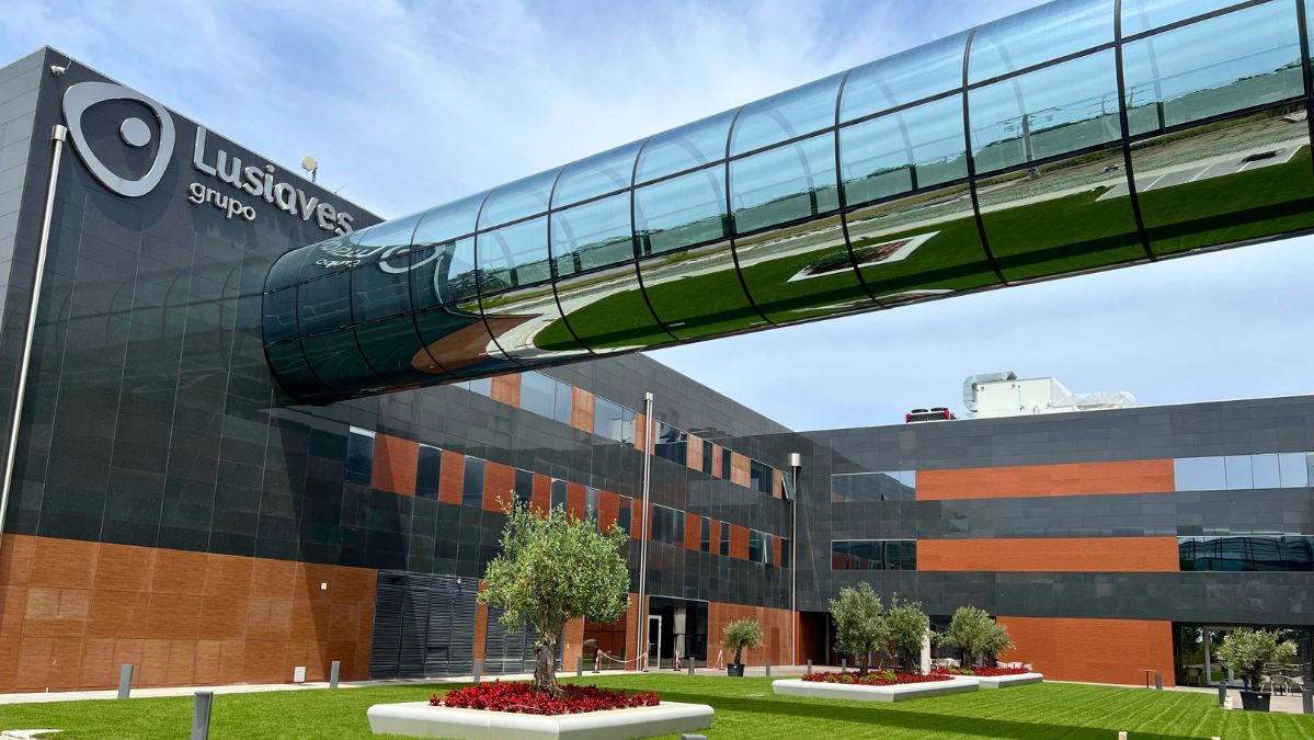 Vista exterior de um edifício com a inscrição «Lusíaves grupo». Uma passagem envidraçada elevada atravessa o espaço sobre uma zona ajardinada.