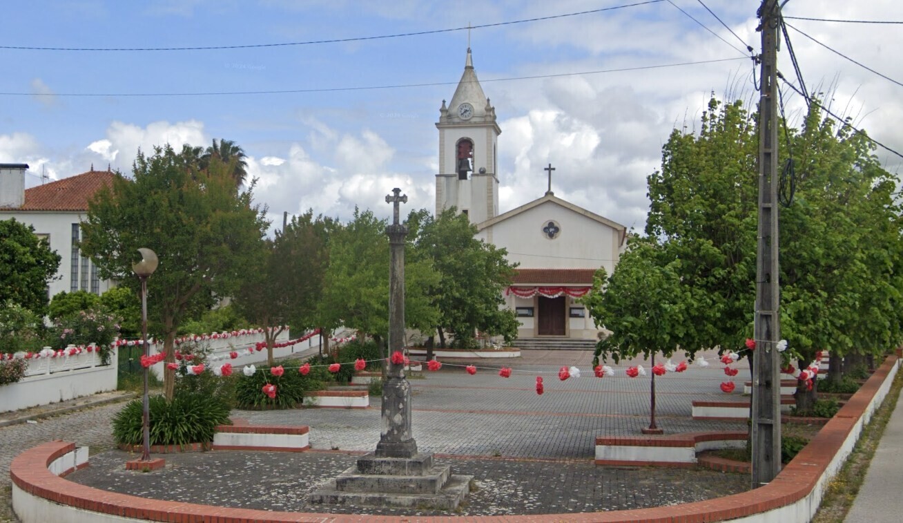 Uma igreja com torre sineira e relógio dá para uma praça arborizada. No centro há um cruzeiro de pedra, e o espaço está decorado com enfeites vermelhos e brancos.