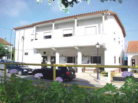 Edifício branco de dois pisos, com telhado de telha e uma entrada coberta. Há automóveis estacionados em frente e vegetação no primeiro plano.