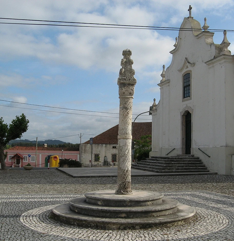 Um pelourinho de pedra ergue-se numa praça empedrada, junto a uma igreja de fachada branca. Ao fundo veem-se outros edifícios e uma paisagem rural.