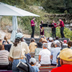 Quatro músicos tocam instrumentos de sopro num palco ao ar livre, perante uma plateia sentada. Há chapéus de sol junto ao público.