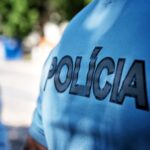 Plano aproximado da farda azul de um agente, com a palavra «POLÍCIA» em destaque. Outro agente surge desfocado ao fundo, num espaço exterior.