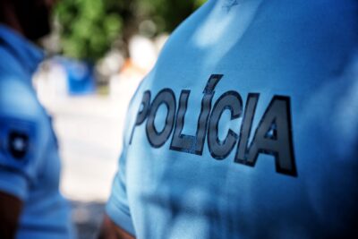 Plano aproximado da farda azul de um agente, com a palavra «POLÍCIA» em destaque. Outro agente surge desfocado ao fundo, num espaço exterior.