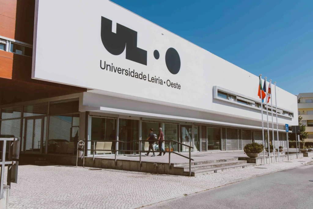 Fachada de um edifício com a inscrição «Universidade Leiria · Oeste». Duas pessoas caminham junto à entrada, onde se veem bandeiras e uma rampa de acesso.