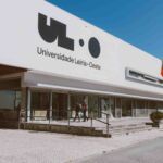 Fachada de um edifício com a inscrição «Universidade Leiria · Oeste». Duas pessoas caminham junto à entrada, onde se veem bandeiras e uma rampa de acesso.