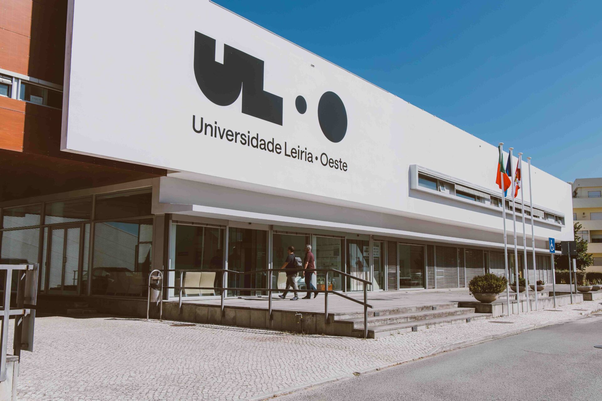 Fachada de um edifício com a inscrição «Universidade Leiria · Oeste». Duas pessoas caminham junto à entrada, onde se veem bandeiras e uma rampa de acesso.