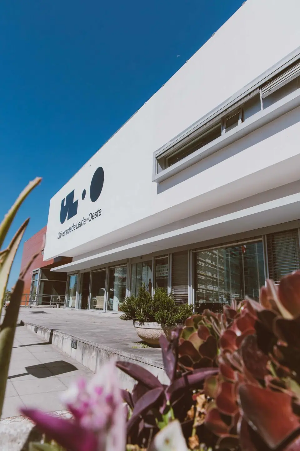 Fachada de um edifício da Universidade Lusófona de Lisboa — Oeste, com o logótipo visível. A entrada envidraçada surge ao fundo, com plantas em primeiro plano.