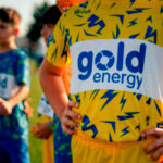 Um grupo de jovens com equipamento desportivo está reunido num campo relvado. Em primeiro plano, uma camisola amarela exibe a inscrição «gold energy».