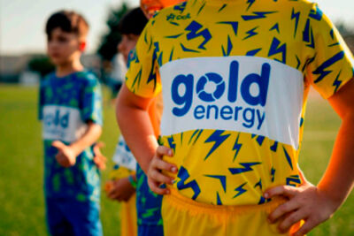 Um grupo de jovens com equipamento desportivo está reunido num campo relvado. Em primeiro plano, uma camisola amarela exibe a inscrição «gold energy».