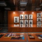 Vista de uma exposição com retratos fotográficos numa parede laranja e publicações expostas em vitrinas. O texto visível não permite identificar com segurança as pessoas retratadas.