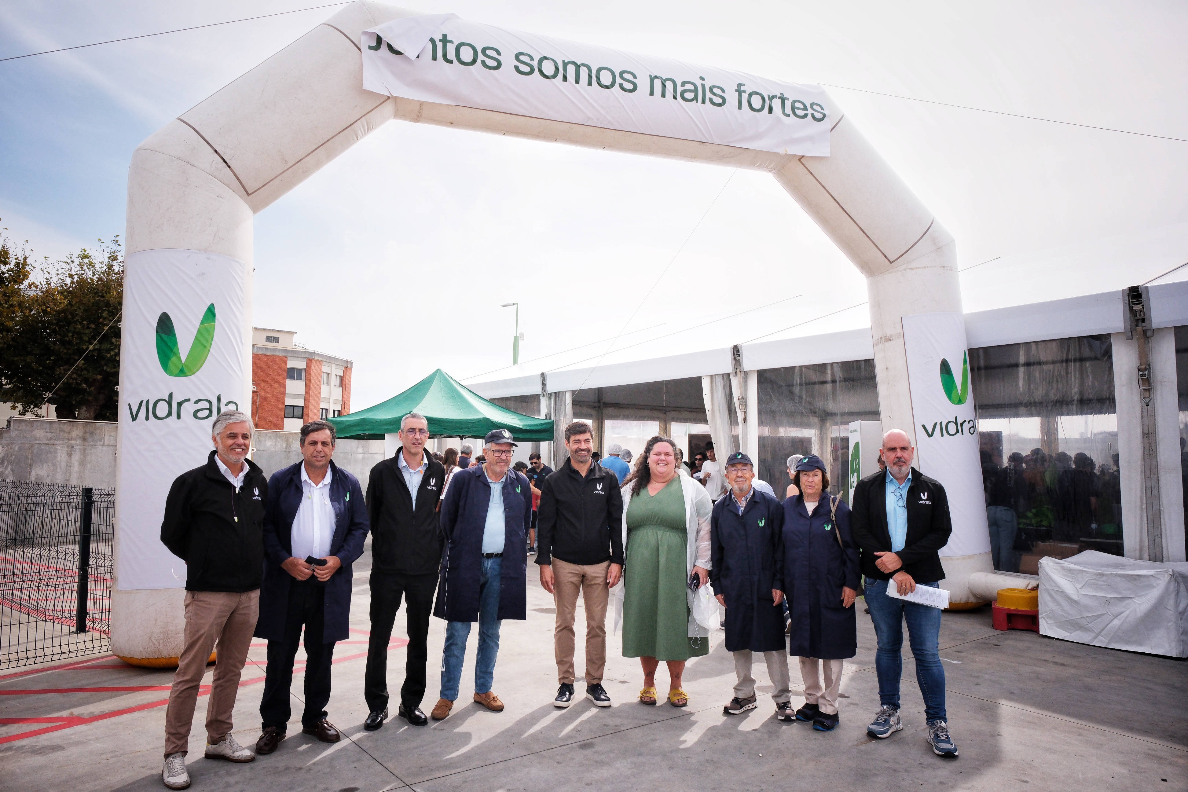 Nove pessoas posam junto à entrada de uma tenda, sob um arco com o logótipo da Vidrala e a frase «Juntos somos mais fortes». Várias vestem roupa com a marca da empresa.