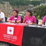 Quatro pessoas participam numa apresentação ao ar livre; uma delas fala ao microfone. Sobre a mesa está uma faixa da Liga Portuguesa Contra o Cancro, Núcleo Regional do Centro.