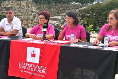 Quatro pessoas participam numa apresentação ao ar livre; uma delas fala ao microfone. Sobre a mesa está uma faixa da Liga Portuguesa Contra o Cancro, Núcleo Regional do Centro.