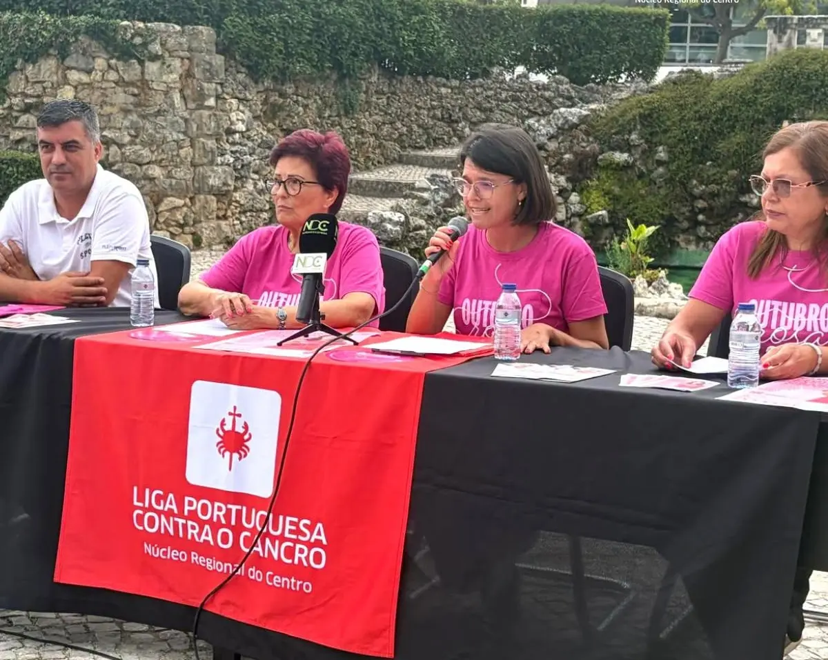 Quatro pessoas participam numa apresentação ao ar livre; uma delas fala ao microfone. Sobre a mesa está uma faixa da Liga Portuguesa Contra o Cancro, Núcleo Regional do Centro.