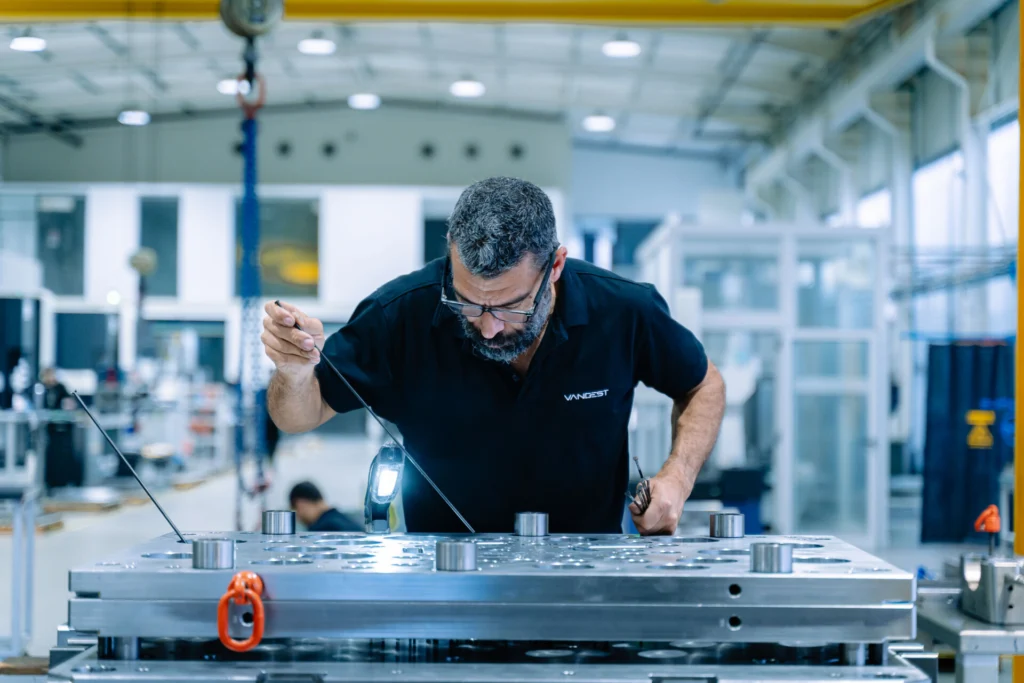 Um trabalhador inspeciona uma grande peça metálica numa oficina industrial. Usa uma luz portátil e instrumentos de medição.