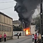 Um incêndio de grandes dimensões provoca chamas e uma coluna de fumo negro numa rua, junto a edifícios. Várias pessoas observam a situação à distância.