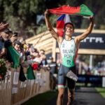 Um atleta português celebra com a bandeira de Portugal erguida ao atravessar uma zona de meta. O público aplaude dos dois lados do percurso.