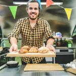 Um homem sorri enquanto apresenta vários pães sobre uma tábua de madeira, atrás de um balcão. Ao fundo, há equipamento de cozinha e bandeirolas coloridas.