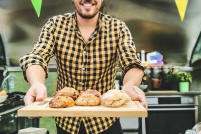 Um homem sorri enquanto apresenta vários pães sobre uma tábua de madeira, atrás de um balcão. Ao fundo, há equipamento de cozinha e bandeirolas coloridas.