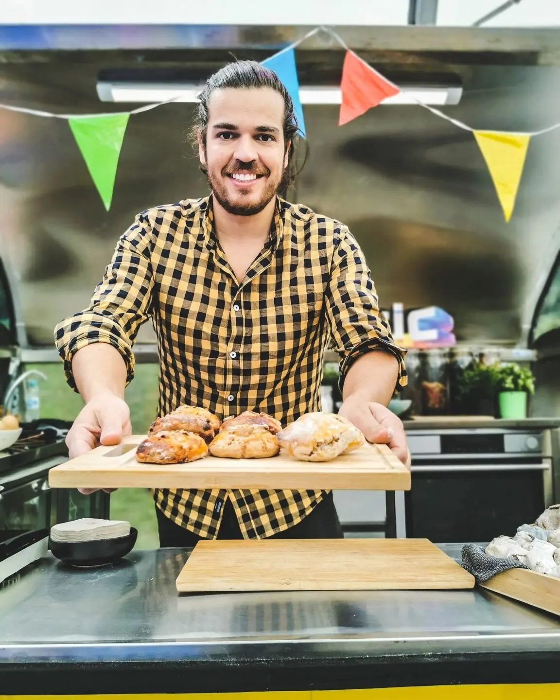 Um homem sorri enquanto apresenta vários pães sobre uma tábua de madeira, atrás de um balcão. Ao fundo, há equipamento de cozinha e bandeirolas coloridas.