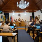 Uma mulher intervém numa reunião numa sala com uma mesa de presidência e vários participantes sentados. Veem-se computadores portáteis, microfones e bandeiras, incluindo a de Portugal e a da União Europeia.