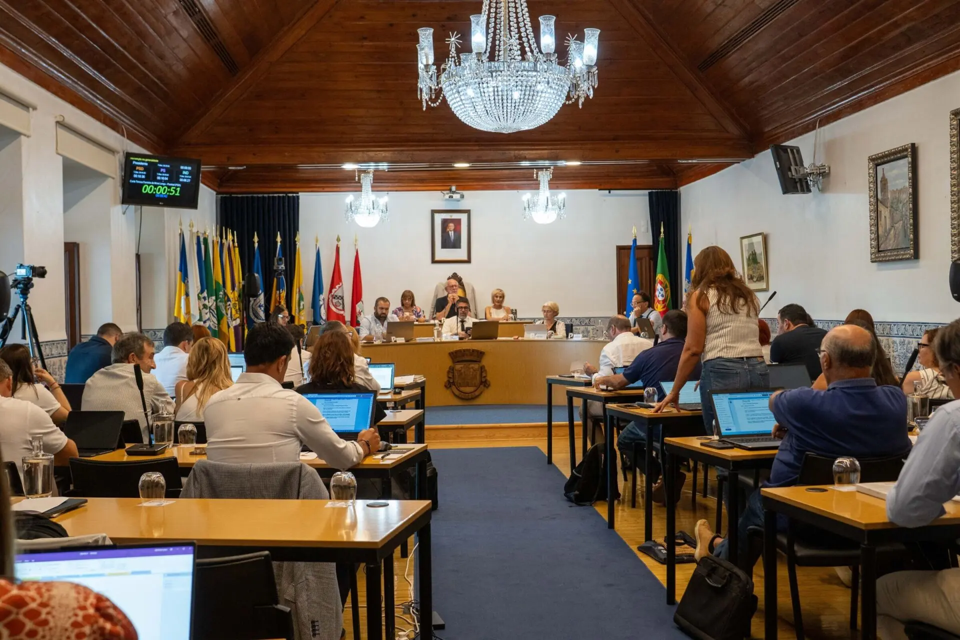 Uma mulher intervém numa reunião numa sala com uma mesa de presidência e vários participantes sentados. Veem-se computadores portáteis, microfones e bandeiras, incluindo a de Portugal e a da União Europeia.