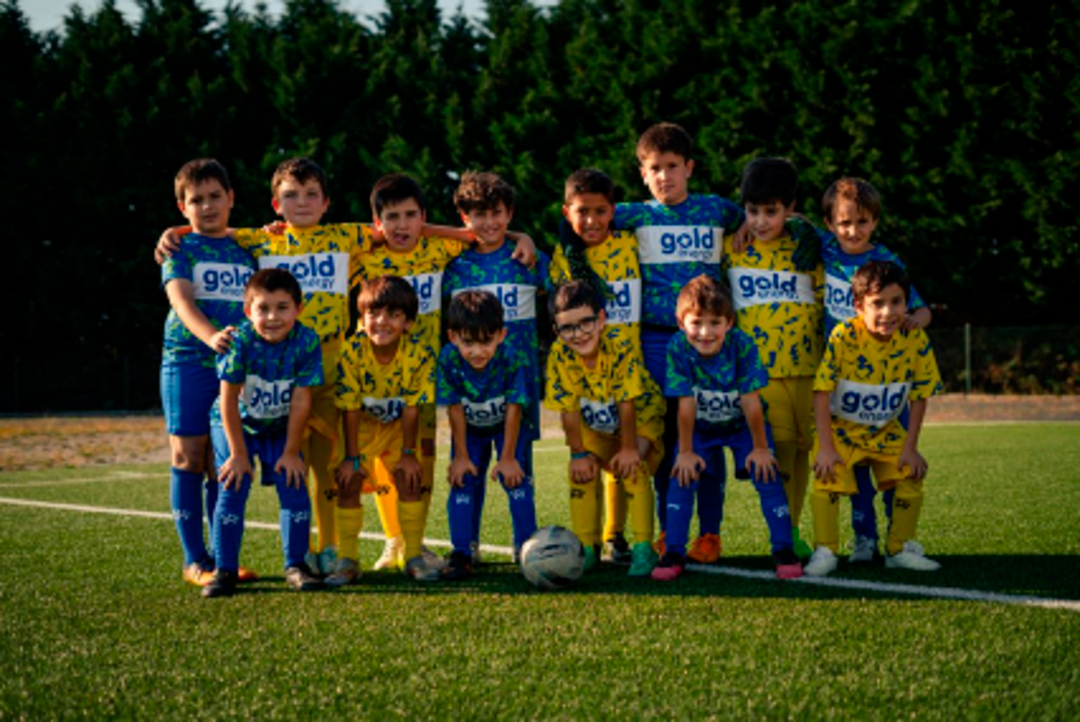 Um grupo de crianças posa em duas filas num campo de futebol, com uma bola à frente. Vestem equipamentos azuis e amarelos.