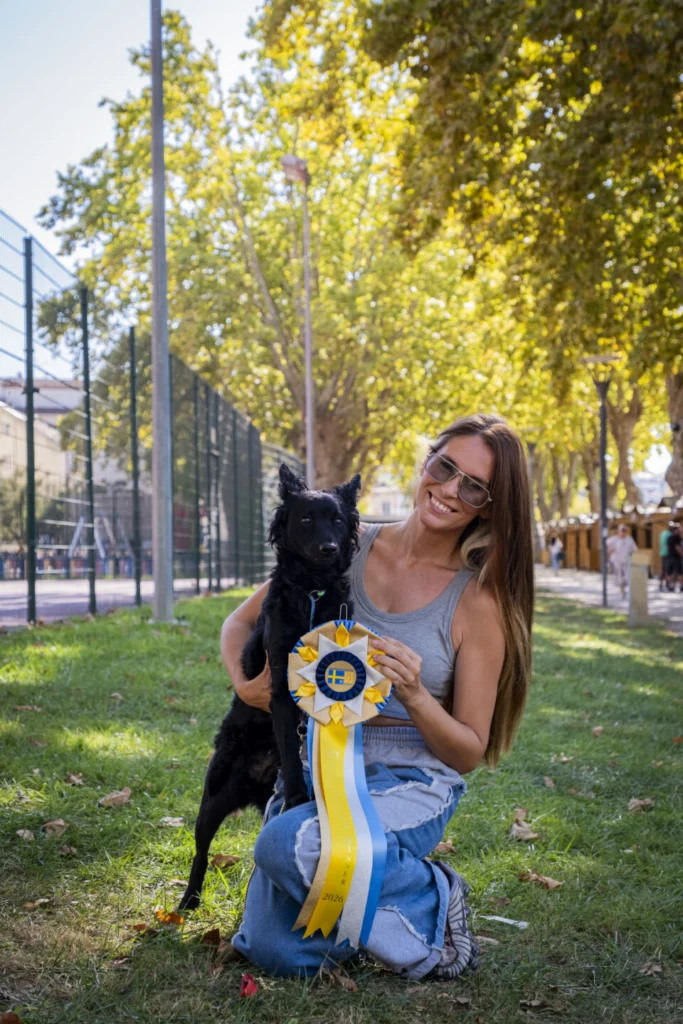 Uma mulher posa com um cão preto num espaço verde junto a um campo vedado. Segura uma roseta azul, amarela e branca, com «2026» visível numa das fitas.