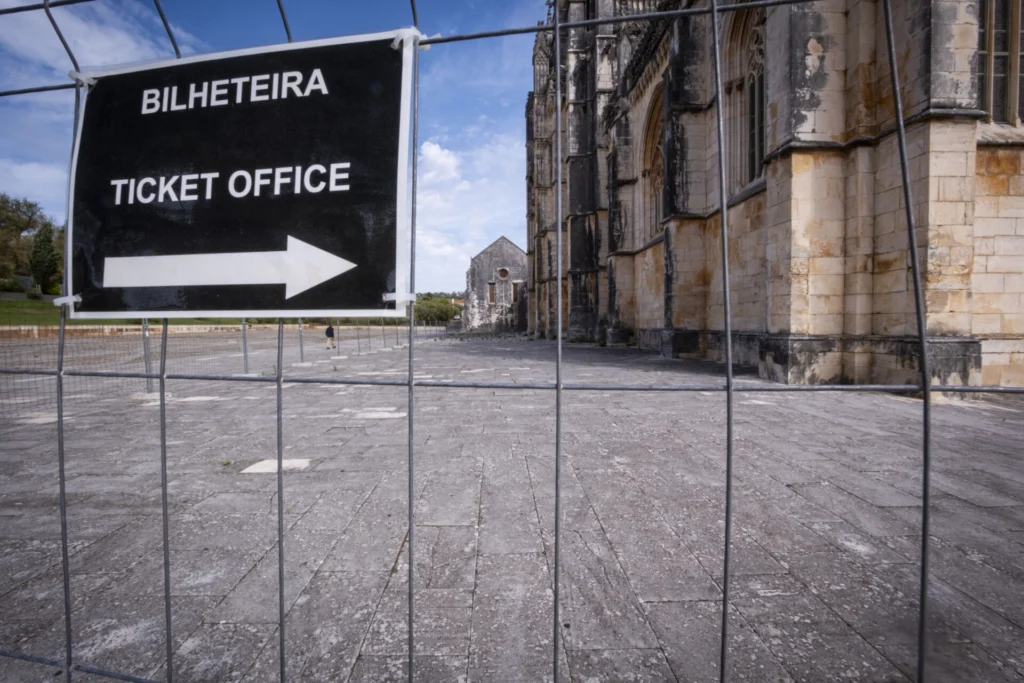 Uma placa com os dizeres «Bilheteira» e «Ticket Office» aponta para a direita. Atrás da vedação, vê-se um edifício de pedra com elementos góticos e uma praça ampla.