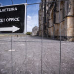 Uma placa com os dizeres «Bilheteira» e «Ticket Office» aponta para a direita. Atrás da vedação, vê-se um edifício de pedra com elementos góticos e uma praça ampla.